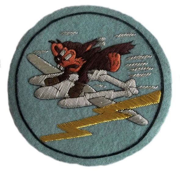 Pale blue circle shaped patch. In center is brown furry cartoon character wearing brown boxing gloves and hanging on to a diving silver plane. Gold lightening flash nearby