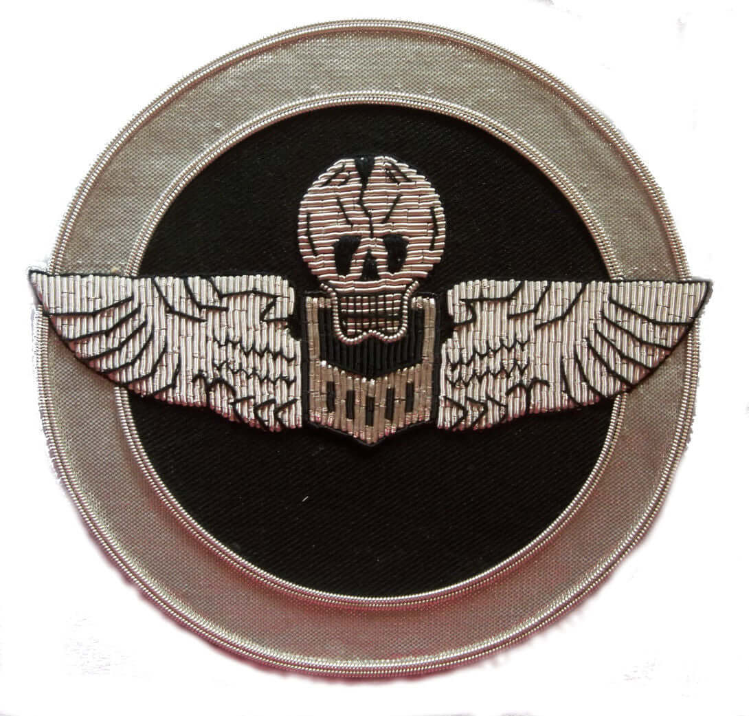 Circle shaped patch with silver wings and skull on black background. Silver outer ring