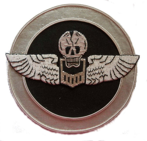 Circle shaped patch with silver wings and skull on black background. Silver outer ring
