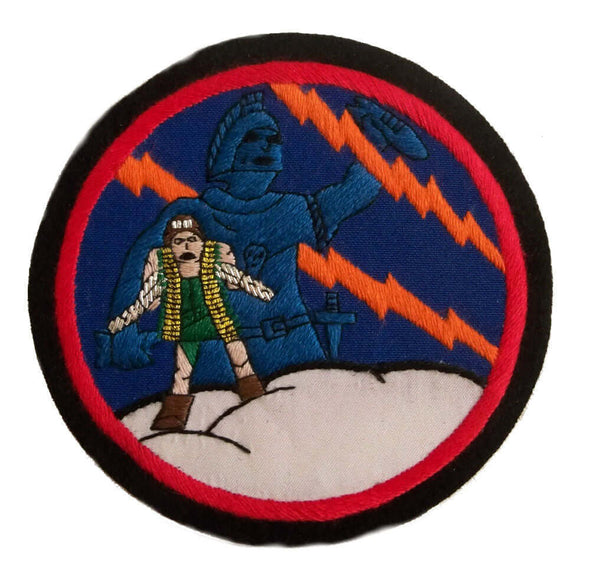 Circle shaped patch with small embroidered man holding two machine guns standing on white cloud,  with large blue giant man behind him.  On dark blue background with red lightening flashes