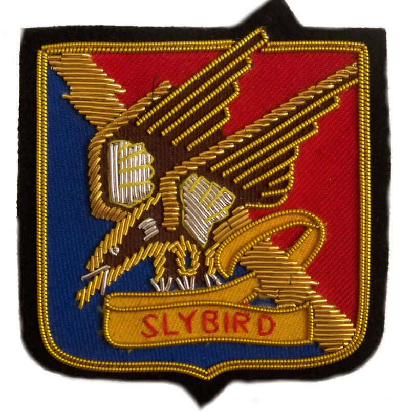 Square shaped patch with gold and silver bird embroidered on blue and red background, with word 'Slybird' under bird