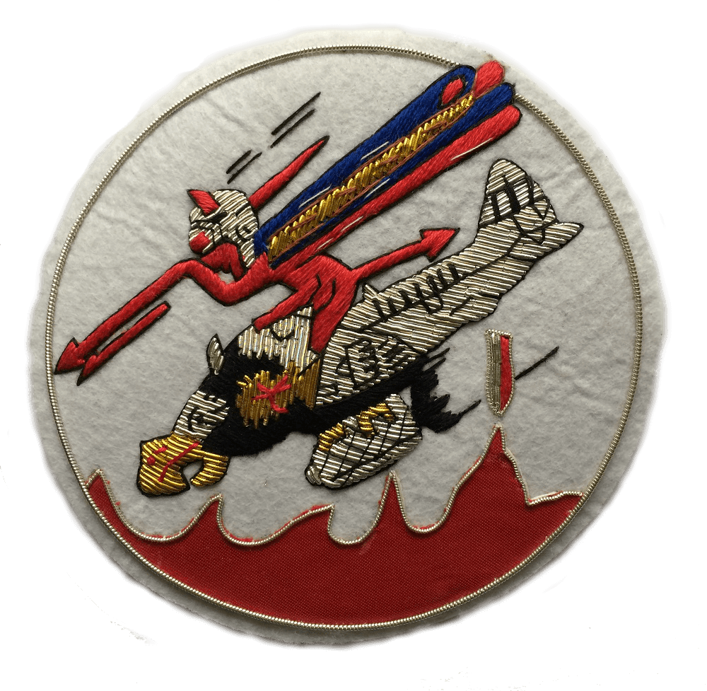 White circle shaped patch.  With Red devil character riding on a plane with a bird's head, and holding a red spear.  Bomb dropping from under the plane.