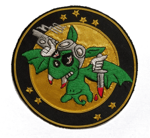 Gold circle shaped patch with black outer ring.  In center is a Green bat, with blood dripping from fangs, holding a blood tipped dagger in left hand and a smoking gun in right.
