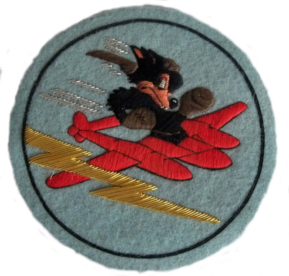 Pale blue circle shaped patch.  In center is a Black wolf character wearing brown boxing gloves riding a red plane.  Gold lightening flashes nearby.