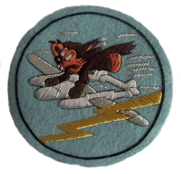 Pale blue circle shaped patch.  In center is brown furry cartoon character wearing brown boxing gloves and hanging on to a diving silver plane.   Gold lightening flash nearby