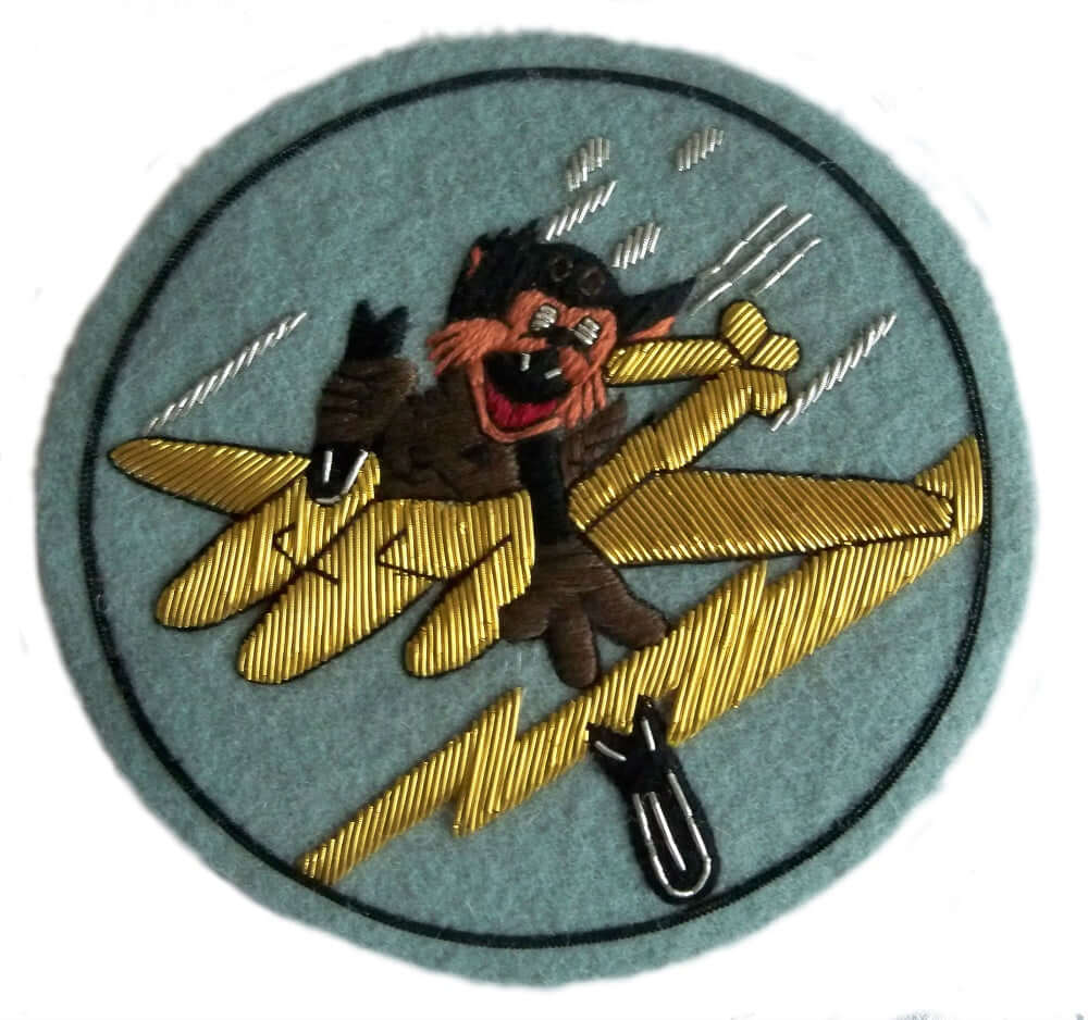 Pale blue circle shaped patch.  In center is brown cartoon character riding on gold plane and throwing bombs from the plane