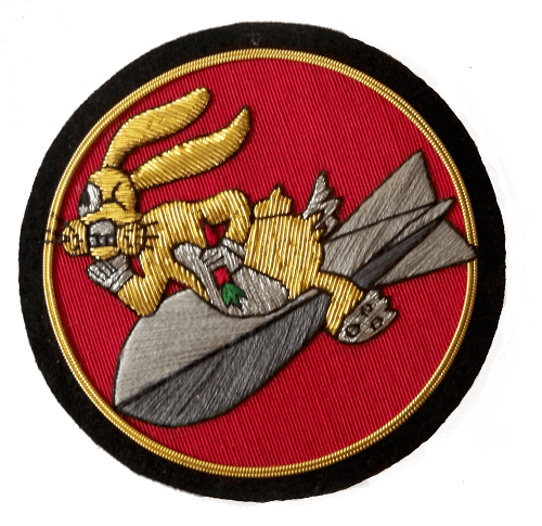 Red circle shaped patch with gold and black outer rings.  In center is a gold colored  'Bugs Bunny' style character, holding a carrot and riding on a grey missile