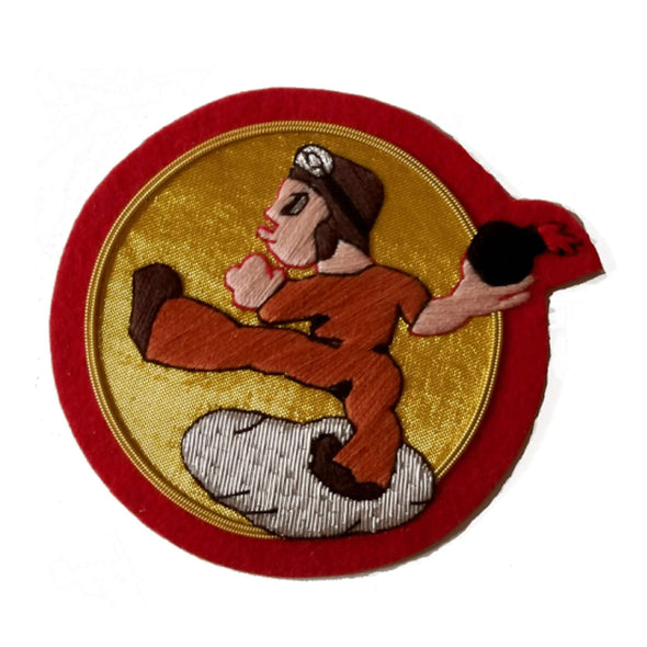 Gold circle shaped patch with red outer ring.  In center is cartoon man wearing a flying helmet and goggles, and throwing a bomb with a lit fuse