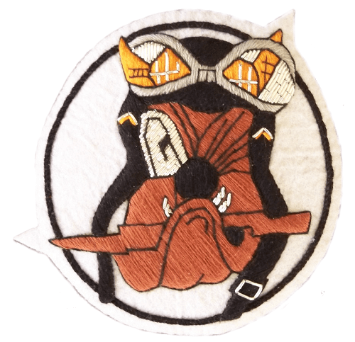 White circle shaped patch with black outer ring.  In center is grumpy bulldog head, holding wooden stick in mouth and wearing a flying helmet and goggles.  His left eye is closed.