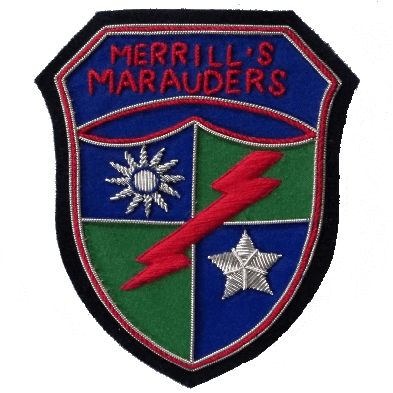 Shield shaped patch with blue and green quarters.  Silver sun in top right quarter, silver star in bottom left quarter.  Red lightening flash runs top left to bottom right.  On top is banner with words, 'Merrill's Marauders'